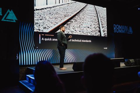 Me on stage at Domain Summit 2026 in Berlin, mid-talk, with a slide reading "A quick anecdote about technical standards" over a close-up of railroad track behind me