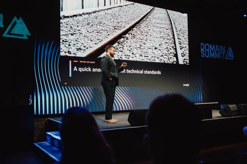 Me on stage at Domain Summit 2026 in Berlin, mid-talk, with a slide reading "A quick anecdote about technical standards" over a close-up of railroad track behind me