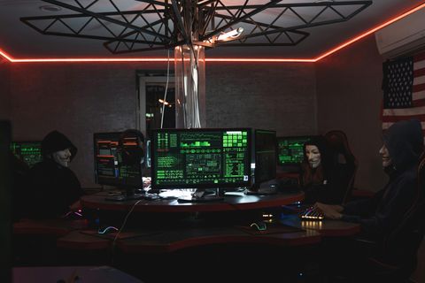 Three figures in Guy Fawkes masks seated at a curved console of triple monitors lit by green hacker-style UI, framed by an industrial geodesic ceiling structure and red rim lighting (photo by Tima Miroshnichenko on Pexels)